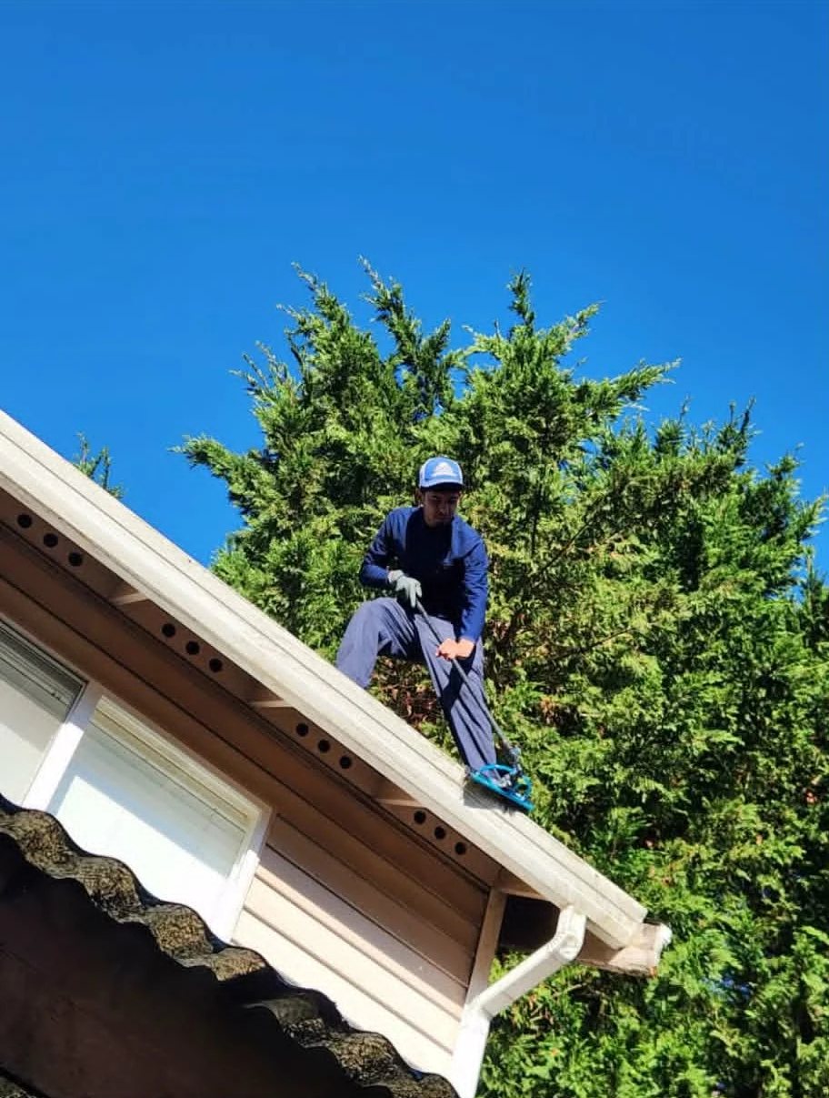 Glen Carrillo, owner of Horizon Glaze, standing on a roof edge, clearing the gutter with a long-handled tool, tall evergreens and blue sky behind