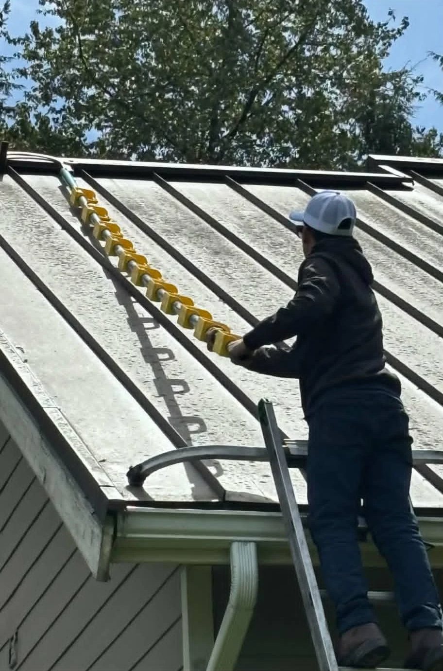 Owner on a ladder rolling a long-handled brush up a metal roof panel